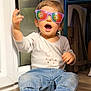 toddler, child, sunglasses, indoor, barefoot, jeans, long_sleeve_shirt, floor, gesture, happy, person, casual_clothing, reflection, flag, purple_sunglasses, home, smile, cute, seated, footwear