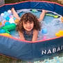 child, curly_hair, pool, inflatable_pool, bubbles, plastic_balls, outdoor, grass, play, smile, toy, summer, water, fun, young, happy, splash, recreation, sunlight, relaxation