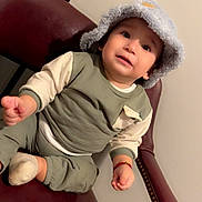Yelimar joined the competition — help win amazing prizes! toddler, child, hat, bucket_hat, sitting, chair, leather_chair, clothing, long_sleeve, pants, footwear, sock, indoor, person, cute, expression, face, hand, curious, cozy
