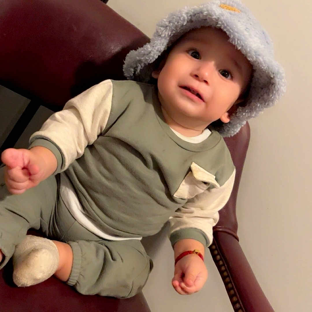 Yelimar joined the competition — help win amazing prizes! bucket_hat, chair, child, clothing, cozy, curious, cute, expression, face, footwear, hand, hat, indoor, leather_chair, long_sleeve, pants, person, sitting, sock, toddler