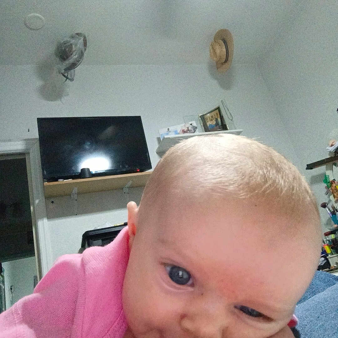 Zyllah is registered to the contest to win money with this photo: baby, infant, pink_clothing, face, blue_eyes, indoor, room, television, hat, shelf, portrait, close_up, skin, child, expression, head, person, furniture, home, casual