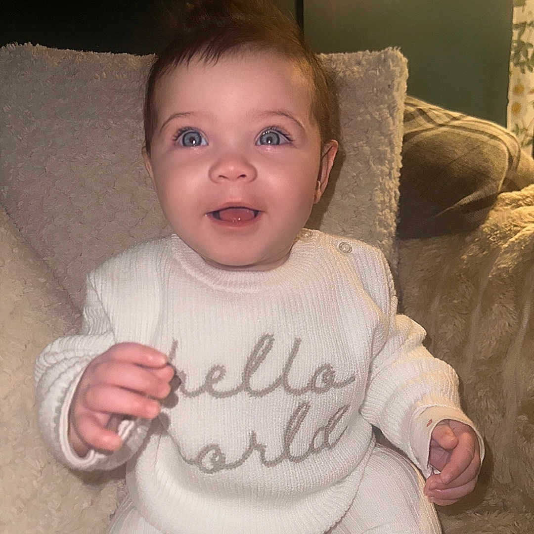 Pixxy is registered to the contest to win money with this photo: baby, blanket, blue_eyes, child, cozy, curious, cute, happy, hello_world, indoor, infant, plush, portrait, seated, small_hands, smiling, soft_lighting, tiny_ponytail, warm, white_sweater