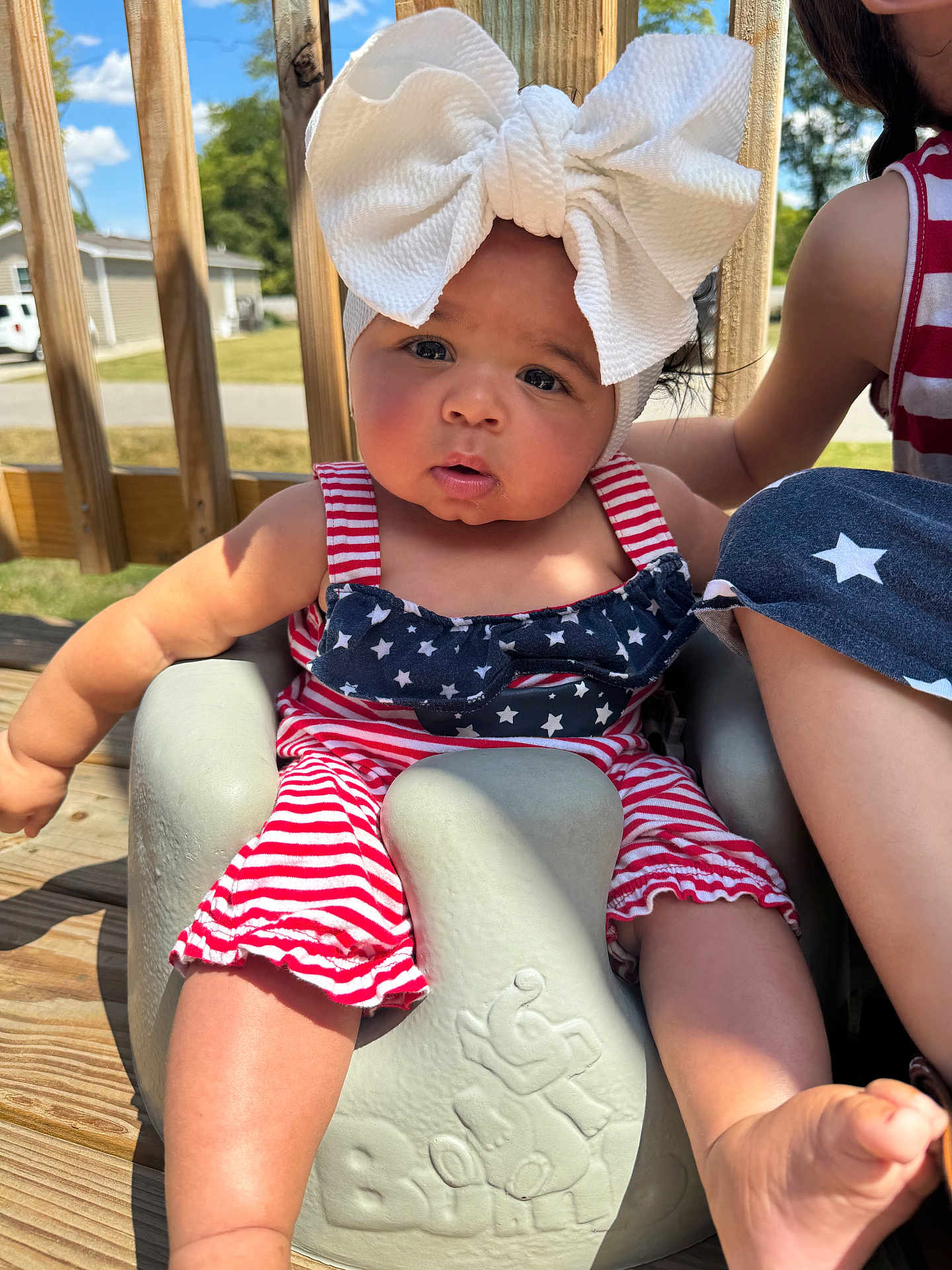 Ocean is registered to the contest to win money with this photo: baby, child, outdoor, sunny, headband, bow, red_stripes, blue_stars, baby_seat, wooden_deck, person, summer, cute, infant, foot, arm, face, expression, clothing, sibling