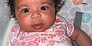 Ocean is registered to the contest to win money with this photo: baby, infant, curly_hair, pink_shirt, floral_pattern, pacifier, cushion, lying_down, wide_eyes, cute, child, person, indoors, closeup, skin, hand, face, expression, young, portrait