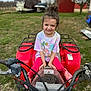 child, girl, smile, outdoor, atv, vehicle, grass, farm, red, clothing, playful, happy, nature, tree, toy, person, casual, daytime, fun, young