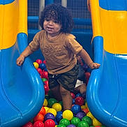Jahmarii is registered to the contest to win money with this photo: child, toddler, curly_hair, ball_pit, slide, blue, yellow, colorful, playground, indoor, fun, play, smiling, clothing, shorts, tshirt, person, hands, legs, happy