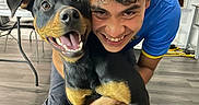 Luna joined the competition — help win amazing prizes! dog, doberman, person, smile, indoor, floor, curly_hair, happy, hugging, table, chair, wall_art, window, pants, blue_shirt, barefoot, wood_floor, home, pet, playful