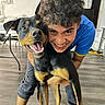 dog, doberman, person, smile, indoor, floor, curly_hair, happy, hugging, table, chair, wall_art, window, pants, blue_shirt, barefoot, wood_floor, home, pet, playful