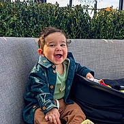 Charbel is registered to the contest to win money with this photo: baby, child, smiling, sitting, sofa, portrait, indoor, chandelier, plants, jacket, clothing, pants, socks, open_mouth, happy, lounge, furniture, window, natural_light, baby_carrier