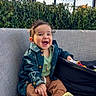 baby, child, smiling, sitting, sofa, portrait, indoor, chandelier, plants, jacket, clothing, pants, socks, open_mouth, happy, lounge, furniture, window, natural_light, baby_carrier