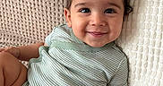 Charbel is registered to the contest to win money with this photo: baby, child, infant, smiling, happy, portrait, indoors, lying_down, blanket, pillow, knit_texture, onesie, green_clothing, brown_hair, face, eyes, arms, legs, cozy, home