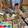 toddler, child, portrait, pouting, sitting, bedroom, bed, blanket, quilt, window, curtains, indoor, educational_book, activity_book, shapes, colors, learning, parent, smartphone, home
