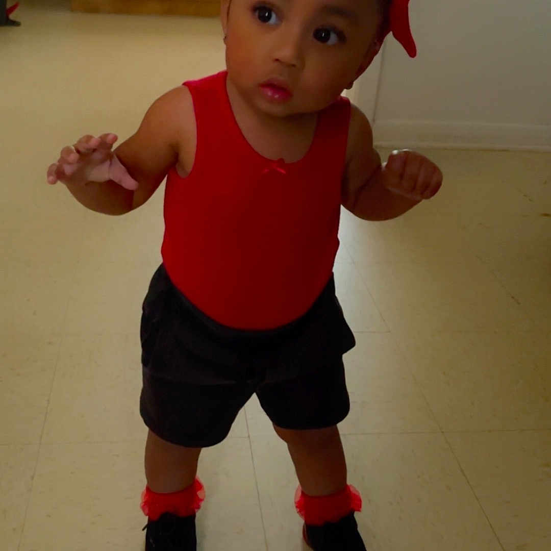 Zaliya Diana is registered to the contest to win money with this photo: baby, black_shoes, cabinet, child, curious, curly_hair, expression, floor, hair_bows, home, indoor, kitchen, microwave, person, red_clothing, shorts, standing, toddler, walking, young_child