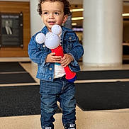 Wander is registered to the contest to win money with this photo: toddler, child, denim_jacket, jeans, curly_hair, toy, black_shoes, indoor, smiling, standing, casual_clothing, converse_shoes, holding_object, earring, floor, background_blur, happy, person, portrait, cute