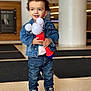 toddler, child, denim_jacket, jeans, curly_hair, toy, black_shoes, indoor, smiling, standing, casual_clothing, converse_shoes, holding_object, earring, floor, background_blur, happy, person, portrait, cute
