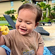 Mailelaulii is registered to the contest to win money with this photo: toddler, child, smiling, flower, outdoor, sunny, brown_shirt, short_sleeves, happy, nature, greenery, lounge_chair, tropical, plant, blue_sky, hair_accessory, cute, portrait, playful, baby