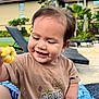 toddler, child, smiling, flower, outdoor, sunny, brown_shirt, short_sleeves, happy, nature, greenery, lounge_chair, tropical, plant, blue_sky, hair_accessory, cute, portrait, playful, baby