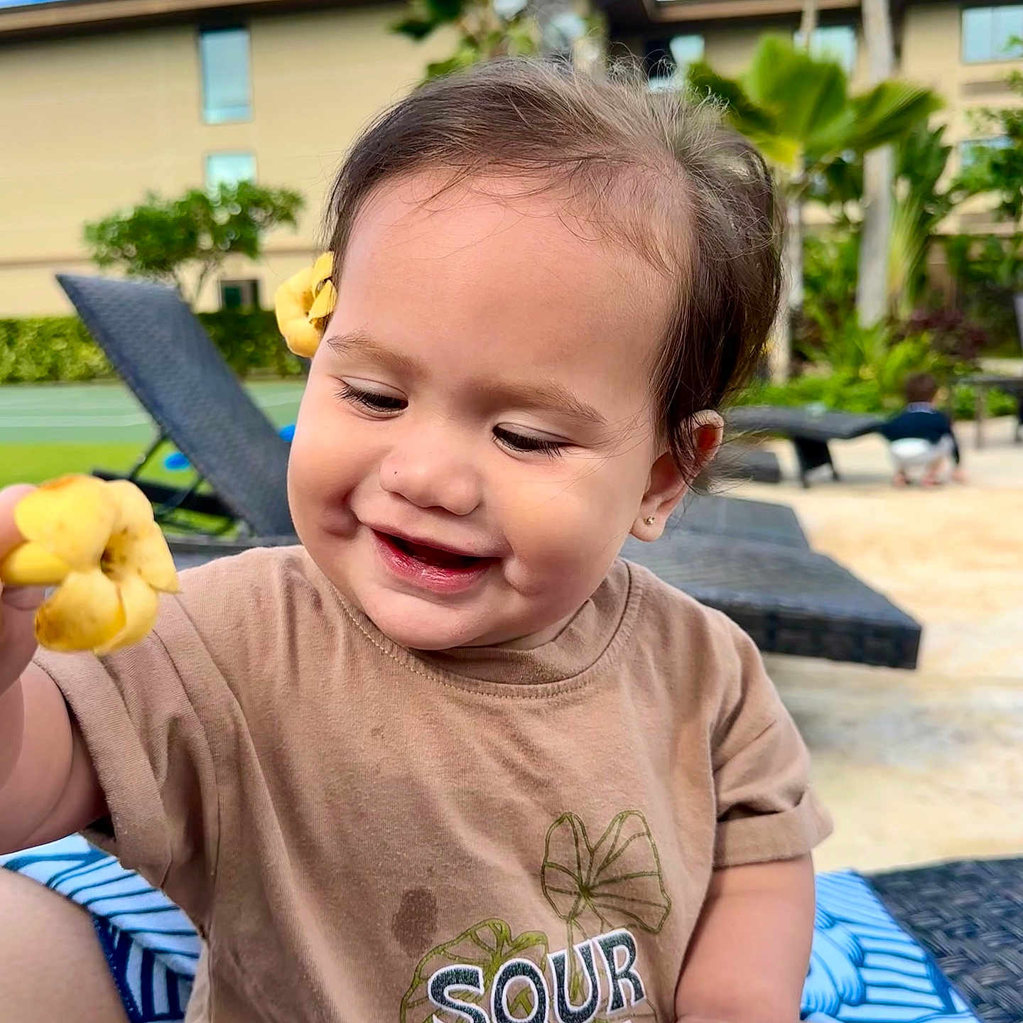 Mailelaulii is registered to the contest to win money with this photo: baby, blue_sky, brown_shirt, child, cute, flower, greenery, hair_accessory, happy, lounge_chair, nature, outdoor, plant, playful, portrait, short_sleeves, smiling, sunny, toddler, tropical