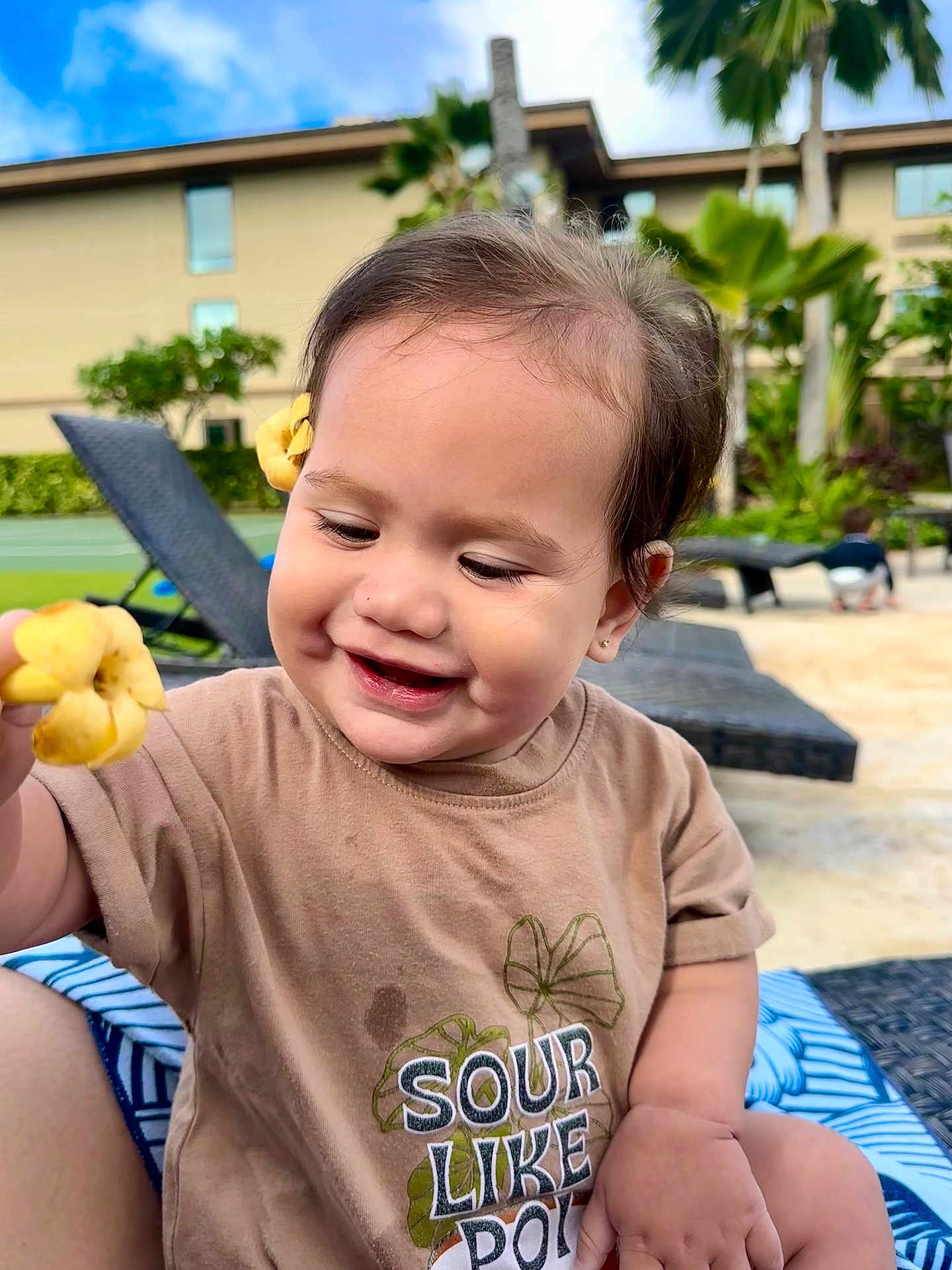 Mailelaulii is registered to the contest to win money with this photo: toddler, child, smiling, flower, outdoor, sunny, brown_shirt, short_sleeves, happy, nature, greenery, lounge_chair, tropical, plant, blue_sky, hair_accessory, cute, portrait, playful, baby