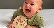 Kannon is registered to the contest to win money with this photo: baby, infant, child, smiling, laughing, portrait, bed, bedding, white_sheets, green_clothing, wooden_sign, milestone, three_months, indoors, close_up, face, hands, happy, sitting, photography