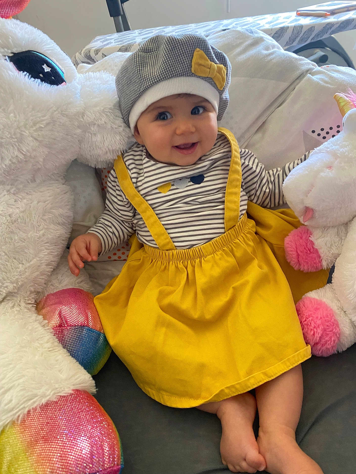 Romy participe au concours pour gagner de l'argent avec cette photo : baby, infant, smiling, beret, hat, yellow_skirt, striped_shirt, stuffed_unicorn, plush_toy, unicorn, soft_toy, cuddly, indoors, pillow, bedlinen, portrait, closeup, cute, hands, feet