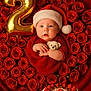 baby, santa_hat, red_outfit, teddy_bear, roses, heart_shape, gold_balloon, number_2, birthday_cake, strawberries, cake, celebration, festive, cute, portrait, infant, holiday_theme, red_background, smiling, child