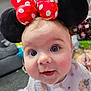 baby, bib, blue_eyes, bow, carpet, child, close_up, curious, face, glasses, indoor, living_room, mickey_mouse_ears, person, playful, polka_dots, red, smiling, sofa, toddler