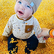 Rodney joined the competition — help win amazing prizes! baby, child, blue_eyes, knitted_hat, corn_kernels, sitting, cute, curious, hand_holding, long_sleeve_shirt, pants, indoor, close_up, infant, soft_lighting, smiling, playful, yellow, warm_clothing, person