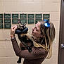 woman, cat, indoor, tortoiseshell_cat, protective_collar, plaque, beige_wall, long_sleeve_shirt, sunglasses, blonde_hair, holding_pet, affection, pet, human, portrait, casual_clothing, bracelet, door, light_switch, expression
