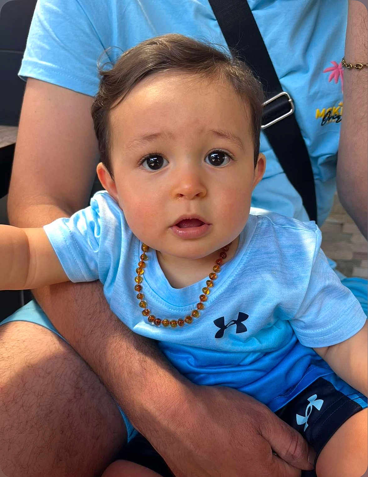 Loann a rejoint le concours — aidez-le/la à gagner de superbes lots ! toddler, child, baby, person, face, eyes, necklace, blue_shirt, adult_arm, holding, casual_clothing, shorts, light_blue, skin, portrait, cute, outdoor, expression, sitting, human