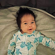 Iyla is registered to the contest to win money with this photo: baby, infant, smiling, face, hair, onesie, turtle_pattern, bed, blanket, lying_down, child, cute, happy, portrait, clothing, young, skin, indoor, person, sleepwear