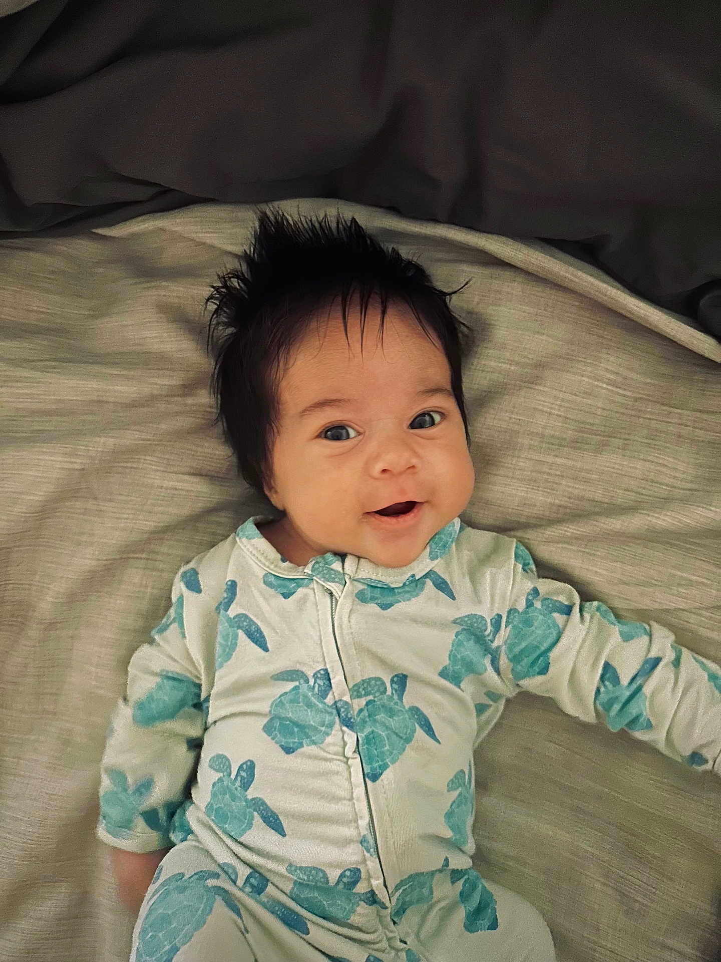 Iyla is registered to the contest to win money with this photo: baby, infant, smiling, face, hair, onesie, turtle_pattern, bed, blanket, lying_down, child, cute, happy, portrait, clothing, young, skin, indoor, person, sleepwear