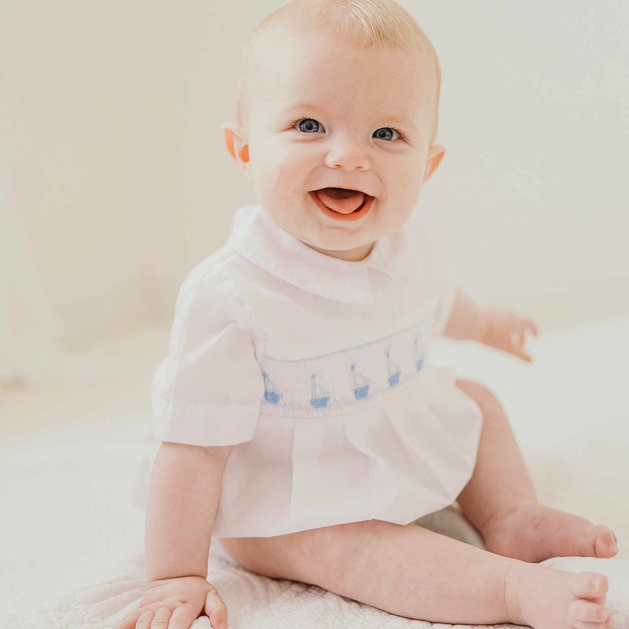 Beau is registered to the contest to win money with this photo: baby, infant, smiling, sitting, white_clothing, blue_eyes, cute, child, portrait, soft_light, quilt, embroidered, happy, innocent, indoor, minimalistic, pale_skin, short_hair, hand, foot