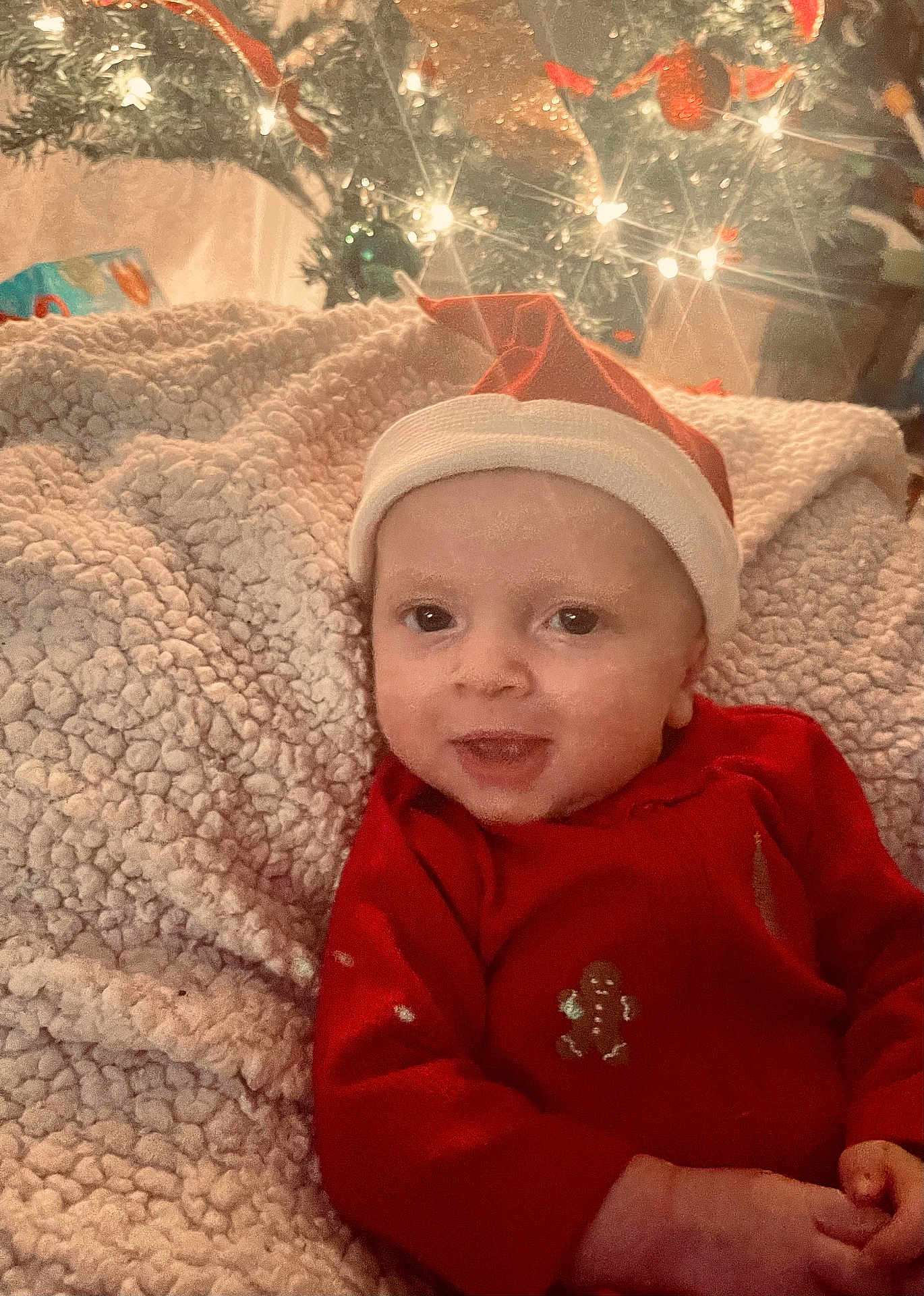 Nicolaos joined the competition — help win amazing prizes! baby, infant, face, smiling, red_clothing, santa_hat, blanket, christmas_tree, holiday, decorations, lights, festive, indoors, cozy, child, portrait, cute, seasonal, happy, celebration