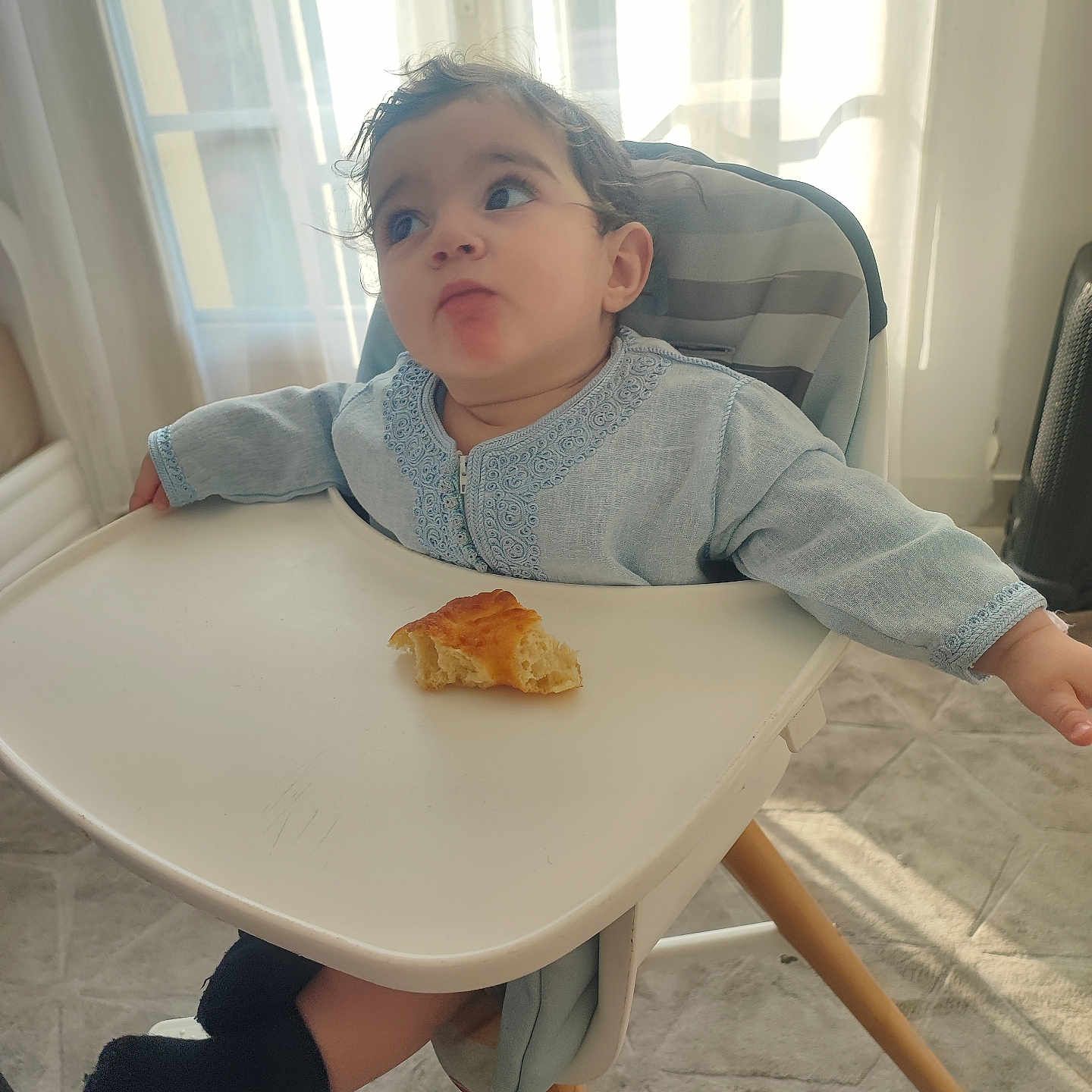 Rayan participe au concours pour gagner de l'argent avec cette photo : toddler, child, high_chair, bread, food, indoor, sunlight, curious, expression, clothing, socks, floor, window, curtains, tray, seat, person, baby, legs, home