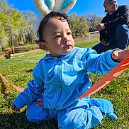 Miller is registered to the contest to win money with this photo: blue_tracksuit, bunny_ears, cap, casual_clothing, child, curly_hair, daylight, family, grass, greenery, kneeling, man, nature, outdoor, park, person, playing, ribbon, sunny, toddler