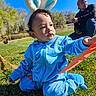 blue_tracksuit, bunny_ears, cap, casual_clothing, child, curly_hair, daylight, family, grass, greenery, kneeling, man, nature, outdoor, park, person, playing, ribbon, sunny, toddler