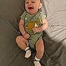 Austin is registered to the contest to win money with this photo: baby, infant, smiling, laughing, onesie, green_clothing, socks, bed, gray_bedspread, happy, cute, child, person, lying_down, indoor, playful, young, portrait, human, casual