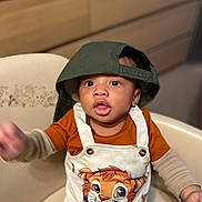 Elozona joined the competition — help win amazing prizes! toddler, child, hat, cap, green_cap, overalls, cartoon_tiger, orange_shirt, beige_chair, indoor, curious, reaching_hand, portrait, cute, baby, clothing, furniture, face, person, young_child