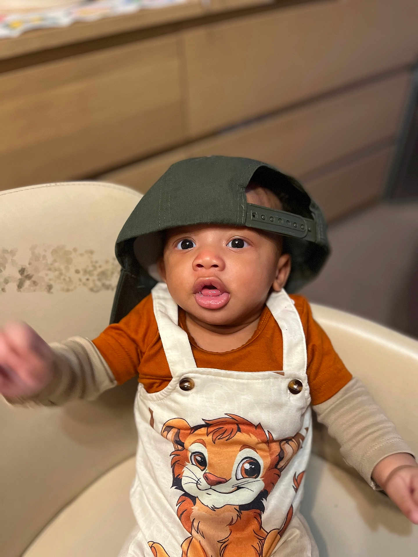 Elozona joined the competition — help win amazing prizes! toddler, child, hat, cap, green_cap, overalls, cartoon_tiger, orange_shirt, beige_chair, indoor, curious, reaching_hand, portrait, cute, baby, clothing, furniture, face, person, young_child