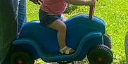 Olivia is registered to the contest to win money with this photo: toddler, child, spring_rider, blue_toy, grass, outdoor, sunlight, playing, person, adult_hands, tattoo, bracelets, shorts, sandals, toy_car, parked_cars, residential_building, grass_field, daytime, greenery