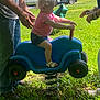 toddler, child, spring_rider, blue_toy, grass, outdoor, sunlight, playing, person, adult_hands, tattoo, bracelets, shorts, sandals, toy_car, parked_cars, residential_building, grass_field, daytime, greenery