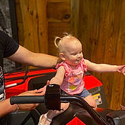 Olivia is registered to the contest to win money with this photo: toddler, child, atv, red_vehicle, wooden_wall, pink_shirt, denim_shorts, sneakers, smiling, arms_outstretched, adult_hand, indoor, playful, hair_tied, person, vehicle_handlebar, supporting_hand, casual_clothing, happy, cute