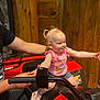 toddler, child, atv, red_vehicle, wooden_wall, pink_shirt, denim_shorts, sneakers, smiling, arms_outstretched, adult_hand, indoor, playful, hair_tied, person, vehicle_handlebar, supporting_hand, casual_clothing, happy, cute