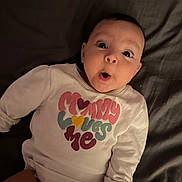 Reagan is registered to the contest to win money with this photo: baby, infant, child, surprised, expression, face, eyes, mouth, clothing, shirt, text, hand, lying_down, blanket, indoor, portrait, cute, adorable, young, person