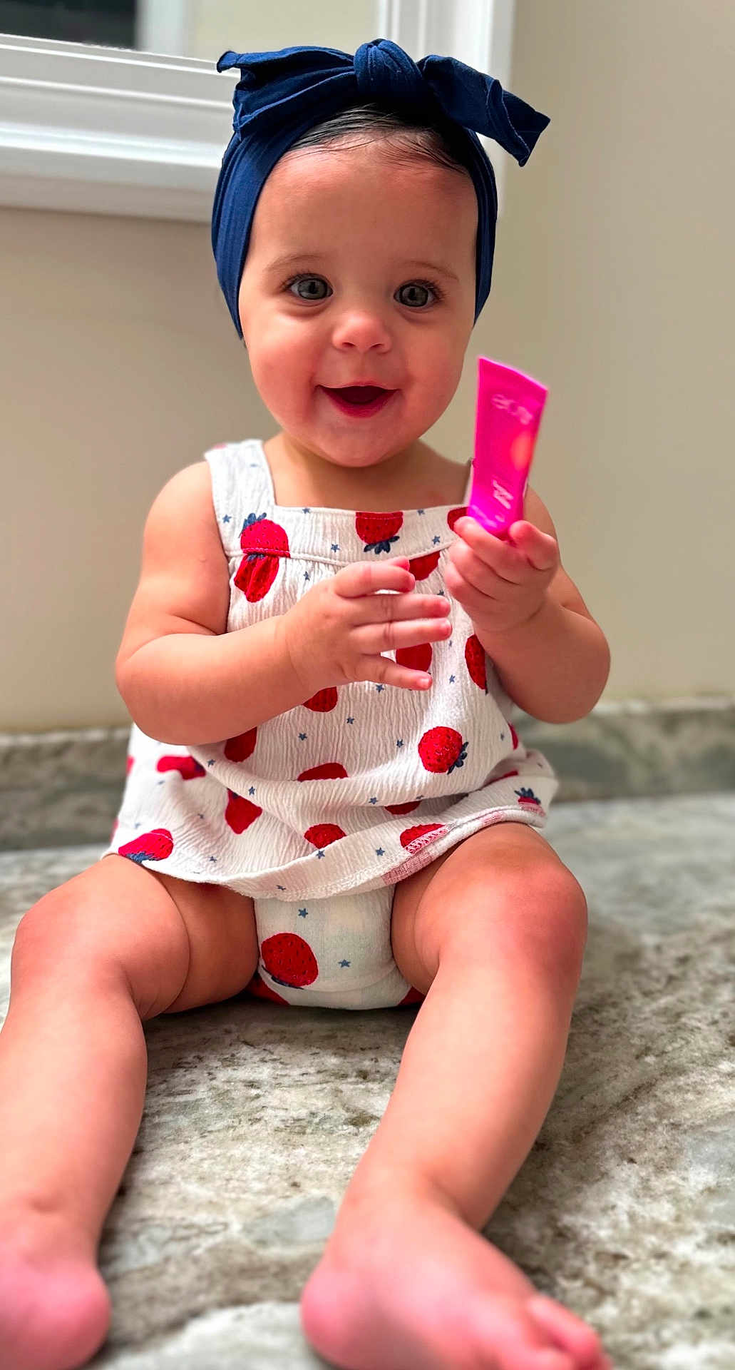 Nora joined the competition — help win amazing prizes! baby, child, smiling, headband, blue_headband, strawberry_pattern, white_outfit, sitting, marble_surface, happy, holding_object, pink_object, cute, infant, indoors, portrait, legs, barefoot, skin, face