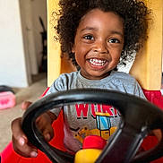 Malakai joined the competition — help win amazing prizes! child, toddler, smiling, curly_hair, toy_car, steering_wheel, indoor, living_room, casual_clothing, happy, playtime, person, young_child, fun, cute, seat, red, yellow, gray_shirt, portrait