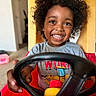 child, toddler, smiling, curly_hair, toy_car, steering_wheel, indoor, living_room, casual_clothing, happy, playtime, person, young_child, fun, cute, seat, red, yellow, gray_shirt, portrait