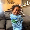 Malakai joined the competition — help win amazing prizes! child, smiling, curly_hair, blue_shirt, dinosaur_graphic, couch, living_room, decorative_plates, yellow_wall, indoor, playful_pose, happy, furniture, cozy, person, portrait, casual_clothing, home, modern_light_fixture, young_child