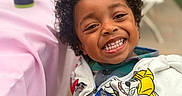 Malakai is registered to the contest to win money with this photo: child, smiling, curly_hair, hoodie, cartoon, happy, face, person, indoor, table, cup, sippy_cup, chair, blurred_background, casual_clothing, close_up, portrait, young_child, playful, seated